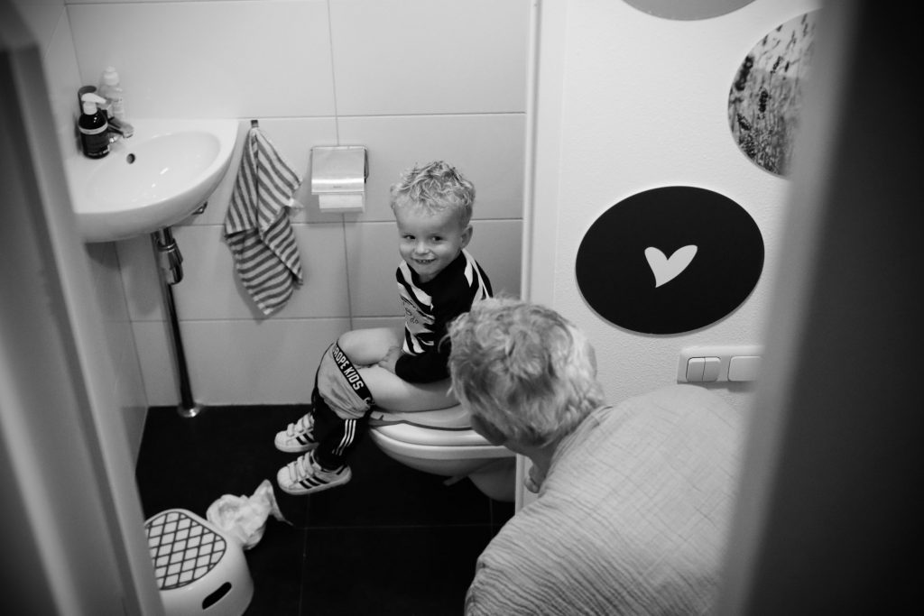 Kleinzoon met blonde krulletjes zit op een moderne WC en Oma komt om de hoek kijken hoe het gaat. Kleinzoon werpt haar een guitige blik toe.