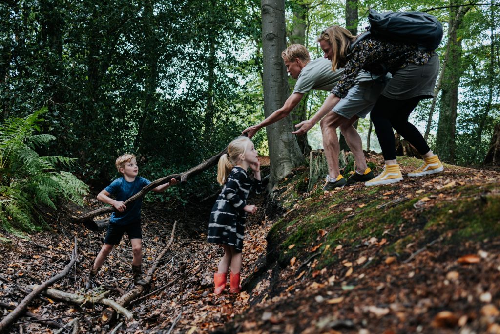 Gezin speelt in het bos: de kinderen zijn een greppel ingesprongen om grote takken te verzamelen voor een hut, maar krijgen hulp van hun ouders om weer uit de greppel te klimmen met het zware hout.