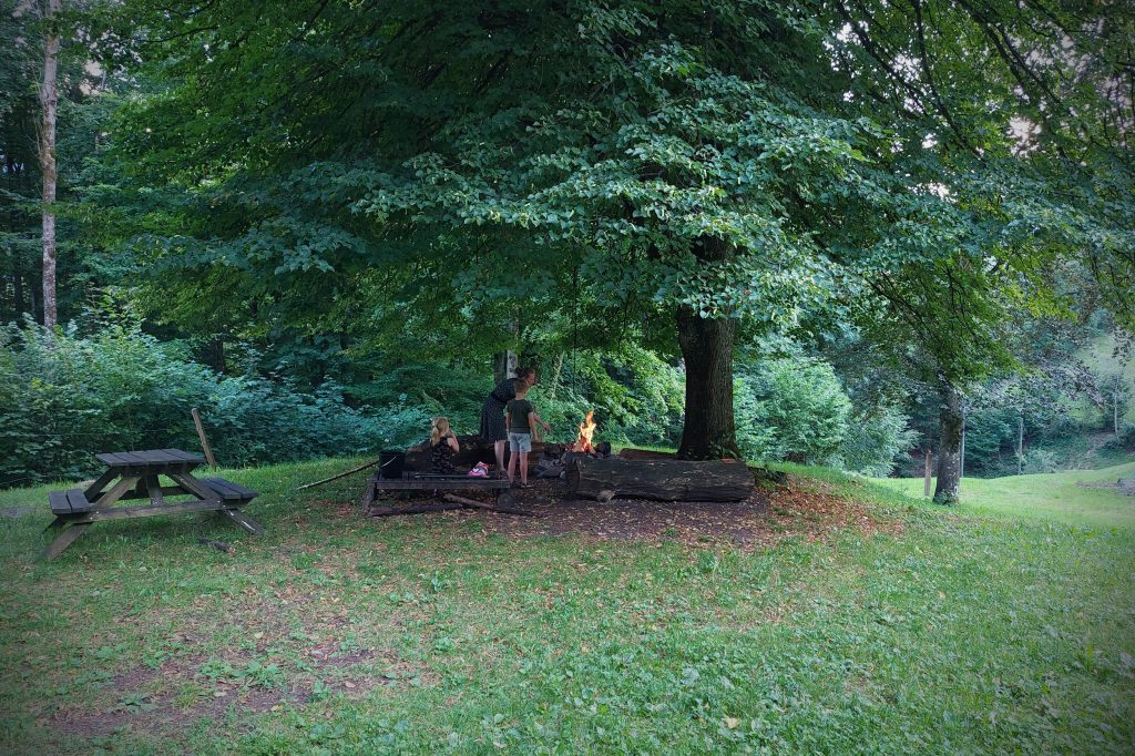In een grote, bosrijke tuin stookt een moeder een kampvuurtje met haar kinderen, bij de vuurplaats onder een grote boom. Het is zomer.