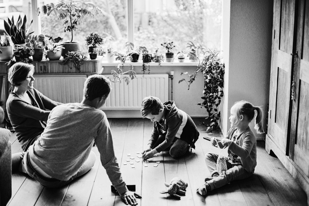 zwart-witfoto van een gezin dat Rummikub speelt op de houten vloer van een huiskamer vol planten.