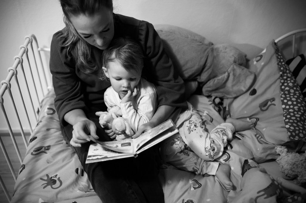 zwart-witfoto van een documentaire gezinsshoot van ellenklikt. moeder leest een boekje voor aan haar peuterdochter, die in haar eenhoornpyjama met fris gewassen haartjes in bed bij haar op schoot ligt.