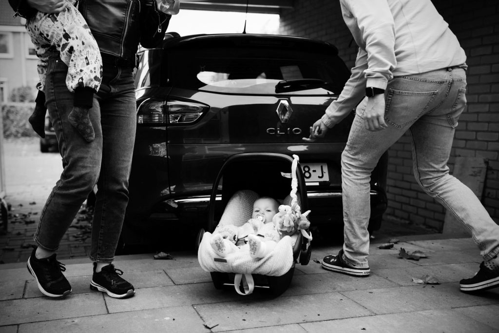 Te midden van tumult bij de auto in- en uitgeladen wordt, zit een baby'tje rustig in de Maxi-Cosi te wachten op de oprit totdat iemand haar naar binnen draagt.