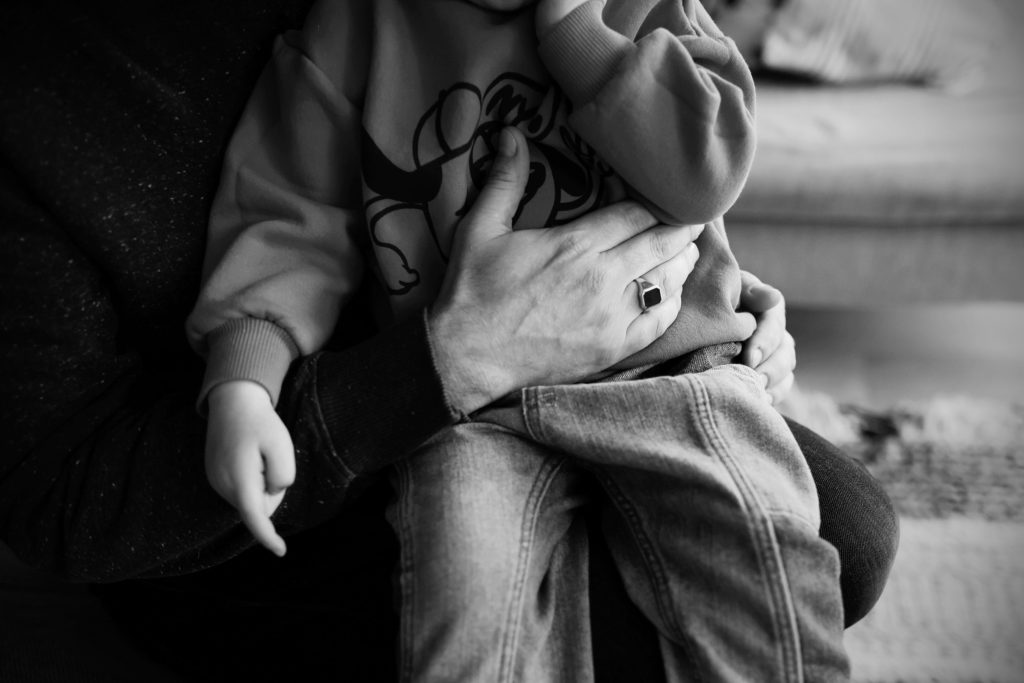 Zoontje krijgt een lieve knuffel van de vader bij wie het op schoot zit. de kidnertrui en spijkerbroek zijn karakteristiek voor het kleine mannetje, de zegelring en donkere trui horen duidelijk bij de vader, die de geborgenheid biedt.